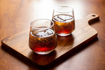Two glasses of dark soda with ice cubes sit on a wooden serving board, placed on a wooden table under warm lighting.
