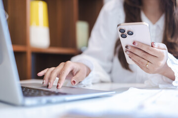 Woman multitasking by using smartphone and typing on laptop at desk, showing focus and productivity...