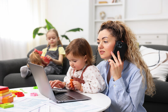 Work-family balance. Single mother holding daughter and talking on smartphone while her other child playing on sofa indoors, selective focus - Powered by Adobe