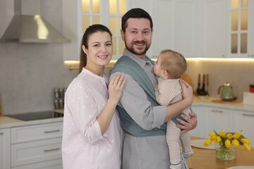 Fototapeta premium Portrait of happy family indoors. Father holding his child in sling (baby carrier) at home