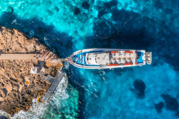 Aerial view of boat with people and rocky sea coast on sunny summer day. Top drone view of yacht, transparent azure water, path. Mediterranean sea. Travel in Sardinia island, Italy. Tropical landscape