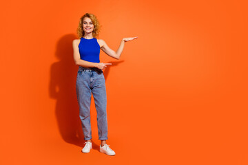 Cheerful young woman with red hair wearing a stylish outfit smiling and gesturing against a bright orange background expressing positivity
