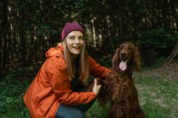 A cheerful young woman wearing an orange jacket and beanie poses with her Irish Setter dog in a...
