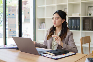 Fototapeta premium Confident businesswoman working on a laptop while reviewing financial documents in a modern office.