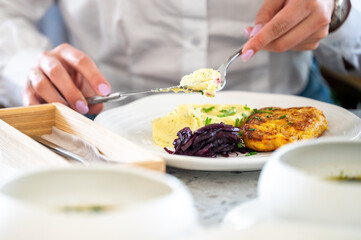 A person is dining, using a fork and knife to eat a meal consisting of mashed potatoes, red cabbage, and a patty. The table setting includes a wooden box with a napkin and utensils.