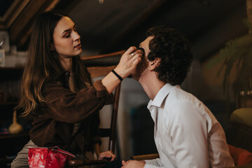 A woman carefully applies makeup to a seated man in a warmly lit room. Both appear engaged in this casual and creative activity, highlighting a blend of artistry and connection in a cozy setting.