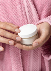 Closeup of woman in pink bathrobe holding cream jar with white lid in hands, cosmetic mockup