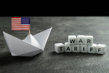 Paper boat with the USA flag and cubes with the text of war tariff on a black background. Concept...
