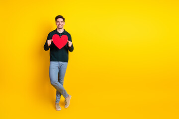Young man in casual green polo holding a red heart shape, smiling cheerfully against a vibrant yellow background.