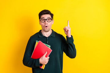 Young man in green polo shirt with book pointing upward on yellow background, expressing excitement...