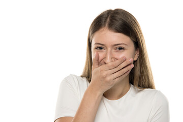 Fototapeta premium Young woman with brown hair covering her mouth with hand while smiling, expressing surprise or amusement against white background