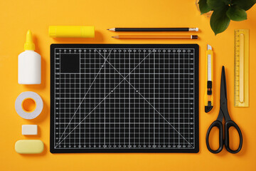 A black cutting mat with white grid lines sits on a bright yellow background surrounded by neatly laid out office supplies: pencils, glue, tape, scissors, cutter, erasers and ruler.