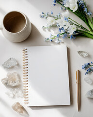 Overhead view of a clean white desk with a spiral notebook, crystals, coffee cup, flowers, and pen arranged beautifully.
