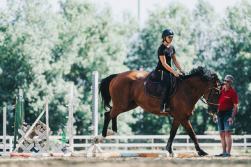 A rider on a horse practices jumping outdoors with guidance from a coach. The scene depicts focus...