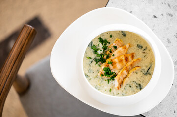 bowl of creamy soup with grilled chicken slices and fresh herbs, served on a white plate. The dish is placed on a stone table with a wooden chair partially visible in the background.