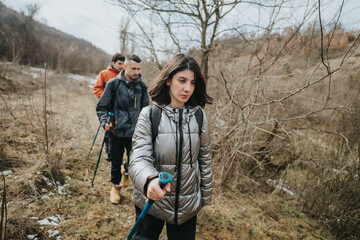 Naklejka premium Three friends trek along a wooded path wearing jackets, enjoying nature during an outdoor hike, with walking poles and a shared adventurous spirit in crisp autumn weather.
