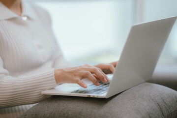 Asian woman living lifestyle wearing wireless headphones staring at laptop screen and communicating with clients freely on sofa at home.
