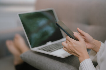 Young Asian woman using phone and computer for video conference while relaxing on sofa at home.
