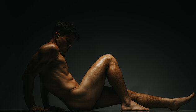An artfully captured image of a male model sitting on the floor, illuminated by moody lighting creating a dynamic and reflective atmosphere, emphasizing form, expression, and aesthetic.