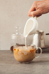 Hand pouring milk into a glass mug with coffee and ice. Cold Brew coffee making process. Vertical background. Copy space for text
