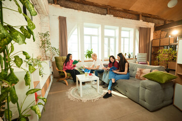 Friends read books together on couch in bright loft interior with plants and coffee table, relaxing in casual atmosphere. Concept of friendship, leisure, lifestyle, communication, learning