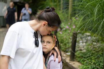 Mom comfortingly hugs her sad daughter in glasses standing in a summer park. Childhood emotions...