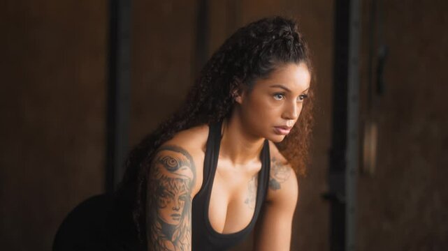 Mid adult Black woman with curly hair performing bent over dumbbell row exercise in dimly lit functional training gym wearing black tank top showing arm tattoo during strength workout