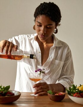 black woman pouring a delightful summer cocktail garnished with flowers and surrounded by fresh fruits.
