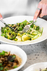 A close-up of hands using a fork and knife to enjoy a fresh salad with cucumber ribbons, leafy greens, and dressing. The image highlights healthy eating with vibrant colors and fresh ingredients