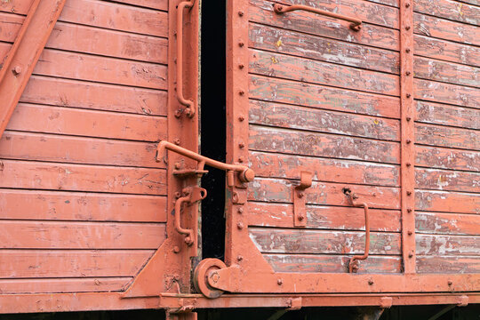 Close-up of an old red wooden freight train door with rusted metal hinges and locking mechanism. Weathered texture and peeling paint highlight the vintage, industrial character. Suitable for themes of