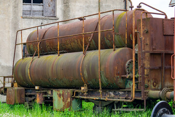 Close-up of heavily rusted cylindrical railway tanks standing next to an abandoned building. Covered with corrosion and moss, the scene captures a sense of industrial decay and the passage of time.