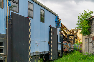 Old blue railway maintenance car with warning markings, positioned on a side track among overgrown grass and decaying buildings. Visible technical equipment and weathered details give the scene an ind
