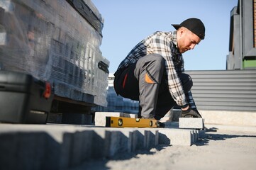 Landscaping paver worker laying paving stones on sandy ground of construction patio yard site in spring summer. Contractor wearing safety protective cloth, gloves for installation work
