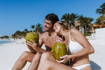 Hispanic young couple holding a coconut and having fun on caribbean beach in holidays or vacations...