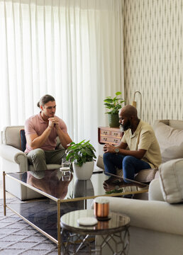Diverse male friends talking in living room at home, with glass coffee table and potted plant