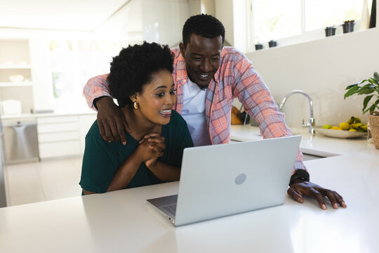 Leaning African American couple looking at silver laptop on kitchen island, with bowl of bananas