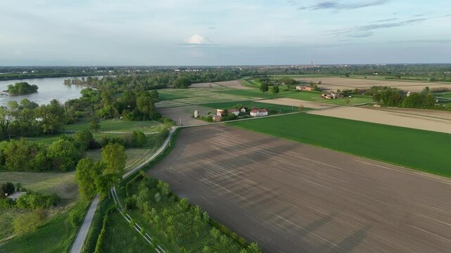 Meandering Po River flows near cultivated fields and patches of forest, distant Cremona skyline with Torrazzo tower visible on horizon under cloudy evening sky, real time drone shot.