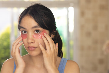 Placing pink hydrogel eye patches under eyes, Asian woman at home near window, wearing blue top