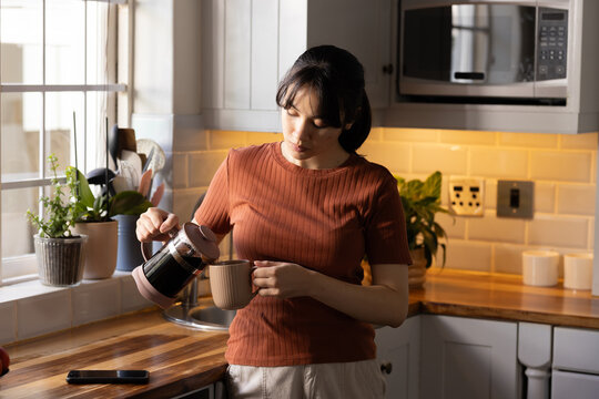 Pouring coffee, Asian woman standing beside countertop in kitchen, with French press and smartphone
