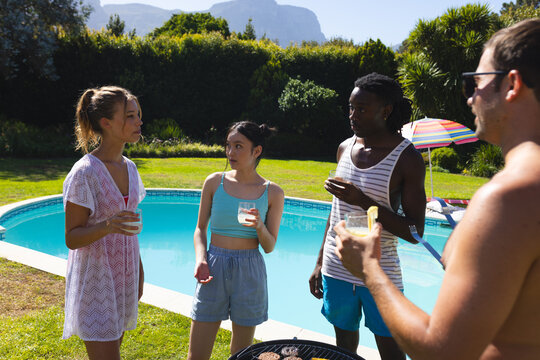 Standing Diverse friends in swimwear holding drinks beside grill by pool, rainbow umbrella