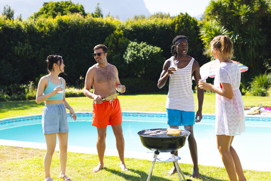 Diverse friends grilling food in swimwear by kidney-shaped pool, with grill and drinks