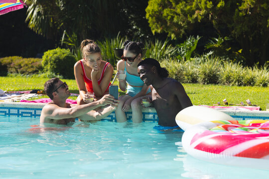 Diverse friends in swimwear taking group selfie at backyard pool, with smartphone and drink glasses