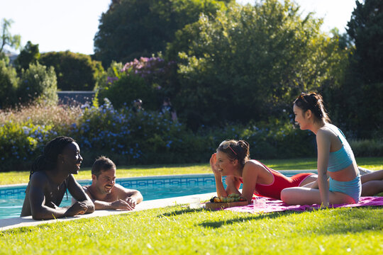 Smiling Diverse friends in swimwear chatting poolside on lawn, with fruit platter on pink towel