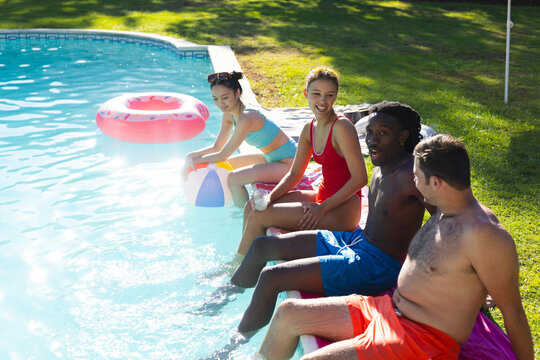 Sitting diverse friends in swimwear dipping legs at backyard pool, with striped beach ball
