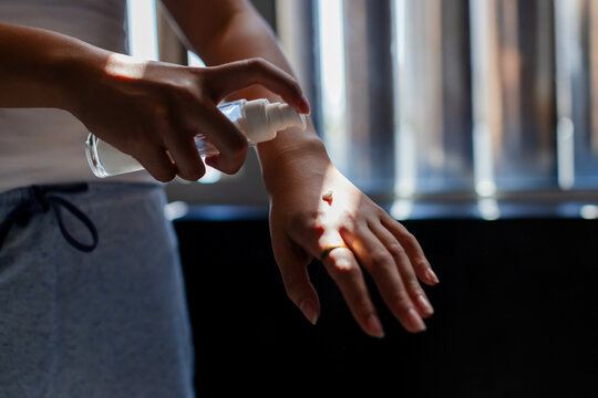 Asian woman dispensing clear liquid onto wrist in room by window blinds, with clear pump bottle