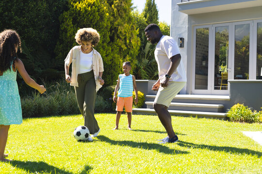 Playing diverse family kicking soccer ball on backyard lawn in front of modern gray house