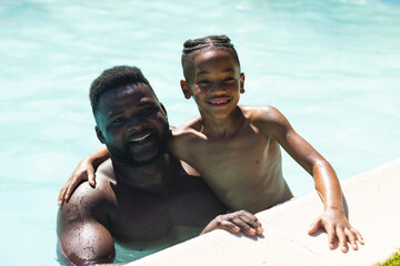 African American father and son leaning on pool edge in outdoor pool, with sunlit water