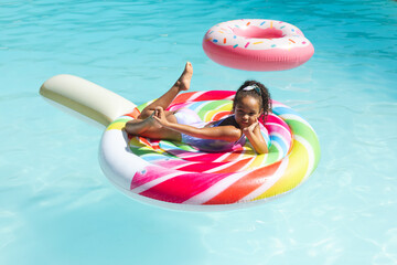 Reclining child girl in swimsuit on rainbow lollipop float in backyard pool, with pink donut float