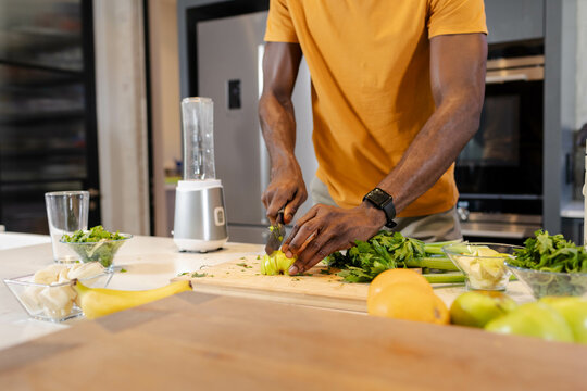 Chopping African American man slicing celery stalks on cutting board in kitchen, with knife blender