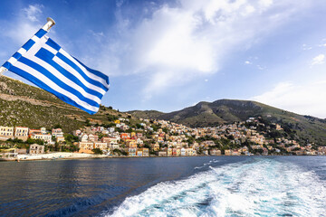 Colorful buildings on a hillside in the idyllic town of Symi by the Aegean Sea on Symi (Simi, Syme) island in Greece on a sunny day. Greek flag is in the foreground.